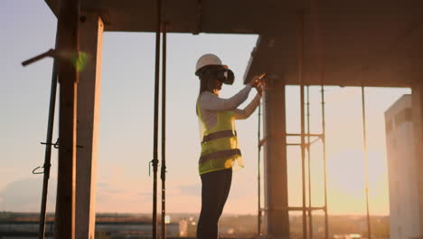 Woman-inspector-in-VR-glasses-and-helmet-checks-the-progress-of-the-construction-of-a-skyscraper-moving-his-hands-at-sunset-visualizing-the-plan-of-the-building